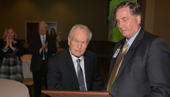 Two men look at a plaque as people in the audience applaud.