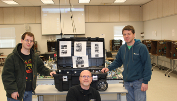 Jim Falbo kneels and two students stand in front of the SWAT robot case