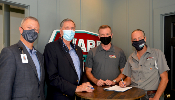 Four men wearing masks stand beside a table.