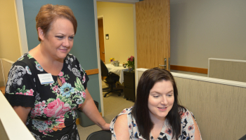 Standing woman guides seated woman through setting up a test on a computer at SCC