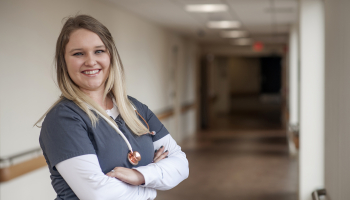 Nurse wearing scrubs and stethoscope stands in hospital hallway.
