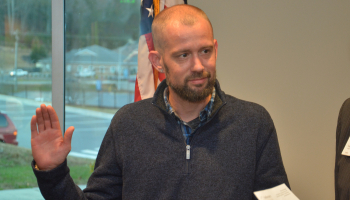 Standing in front of a U.S. flag, a man holds up one hand and places the other on the Bible.