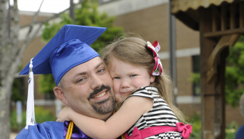 A father, wearing SCC blue cap and gown, holds his daughter outside the college's auditorium.