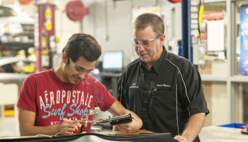 Young man in safety glasses solders two wires together alongside his instructor.