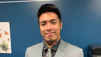 A young man smiles in a gray suit with a floral shirt and silver tie