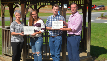 Two young students are holding scholarship certificates in front of a gazebo