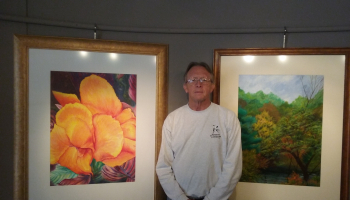Randy Lanier standing between two paintings one is a flower the other a tree scene