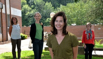 A group of four women stand safely apart while smiling outside