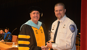 A male college president in a cap and gown presents an award to a male graduate from the emergency medical science program