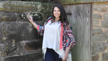 A young woman stands outside of an old wooden building surrounded by nature.