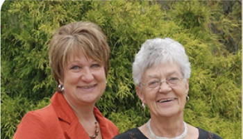 Two women smile and stand side by side outside.