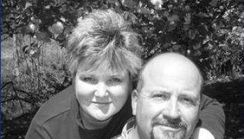 A woman lovingly leans over her husband with her hands placed on his shoulders.