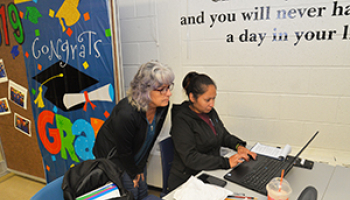 Female instructor leaning over a desk to help female student on a computer.
