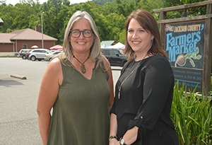 Two female business leaders smile in front of a farmer's market sign on a summer day.