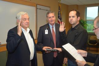 Two men hold up their right hands and place left hands on bible held by a third man. A fourth man reads pledge from a sheet of paper.