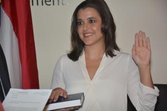 Woman places one hand on a bible and holds the other arm in the air as she's sworn in. Man holds the bible for her.