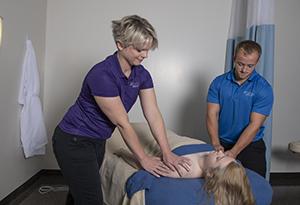 A male and female student gently massage a young woman laying face up on a table.