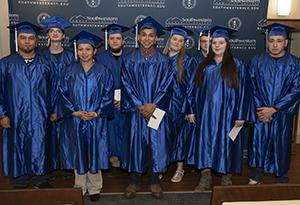 A group of students in blue caps and gowns smile as they sit and wait to receive their diplomas.